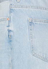 Tessuto in denim azzurro chiaro, caratterizzato da una texture asciugata, leggere sfumature e una tasca posteriore visibile con dettagli in hardware arancione.
