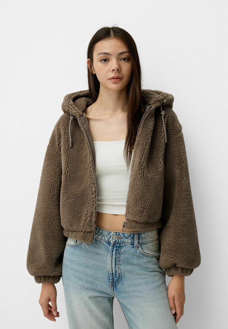 Veste en polaire marron et duveteuse avec capuche, fermeture éclair à l'avant et poignets élastiques, associée à un jean taille haute bleu clair.