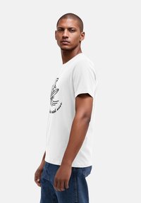 Weißes Baumwoll-T-Shirt mit grafischem Design, das einen Hund auf einem Surfbrett zeigt, mit dem Text "Fang die nächste Welle". Kurze Ärmel, lässige Passform.