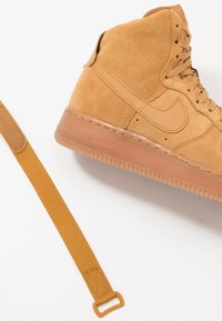 Tan suede high-top sneaker with rubber outsole and signature Nike logo. Accompanied by a matching strap with a rectangular buckle.