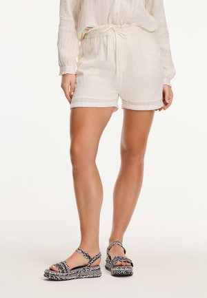 Shorts color crema realizzati in tessuto testurizzato con una vita elastica. Abbinati a sandali intrecciati bianchi e neri con fasce incrociate.