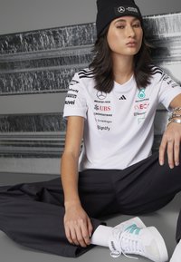 Camisa polo blanca con detalles y logotipos en negro, combinada con pantalones negros y zapatillas blancas con rayas en teal. Muffler negro llevado en la cabeza.