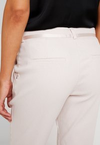 Pantalons taillés rose clair avec une finition texturée, dotés d'une ceinture en satin et de poches arrière décoratives.