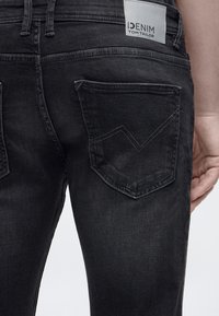 Πίσω όψη ατόμου που φοράει σκουρόχρωμα γκρι τζιν με ραμμένες πίσω τσέπες και μια λευκή ετικέτα Tom Tailor.