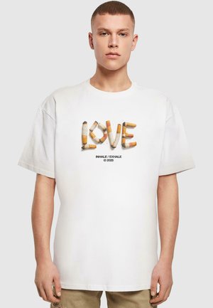 T-shirt en coton blanc avec le mot "AMOUR" composé de mégots de cigarettes. Comprend le texte "INHALE / EXHALE © 2025" sous le design.