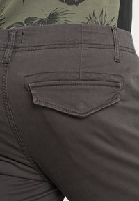 Graue dehnbare Stoffhose mit einer diamantförmigen Gesäßtasche. Die Hose hat eine glatte Textur und dezente Nahtdetails.