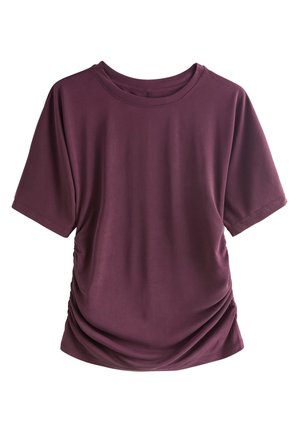 Kvinde med brun hestehale iført en løs mørkelilla top og mønstrede leggings, stående med front mod en lysegrå baggrund.