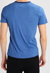 Mężczyzna ubrany w gładką niebieską koszulkę z krótkim rękawem i czarne spodnie, pokazany od tyłu na tle jasnym.