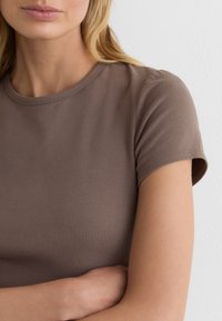 Gros plan d'une femme croisant les bras, portant une chemise marron à manches courtes avec des cheveux blonds ondulés sur l'épaule.