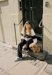 Jeune femme en vêtements décontractés et casquette assise sur le seuil en pierre, souriante, tenant une tasse de café, caressant un petit chien fluffy à ses côtés.
