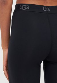 Juodos UGG leggingai, pagaminti iš lygaus, tampaus audinio, su plačia juosta, ant kurios yra UGG logotipas pilka spalva.