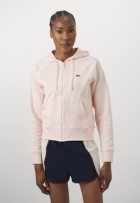 Svijetlo ružičasta zip-up dukserica s kengurudžepom, rebrastim manžetama i malim zelenim logom. Uparen s mornarsko plavim i bijelim šortsem s rubom.