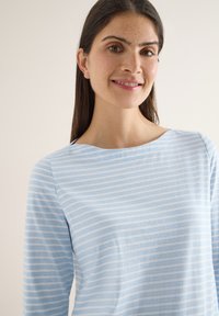 Femme aux longs cheveux noirs et taches de rousseur souriante, portant un chemisier à rayures horizontales bleu clair et blanc, sur un fond uni.