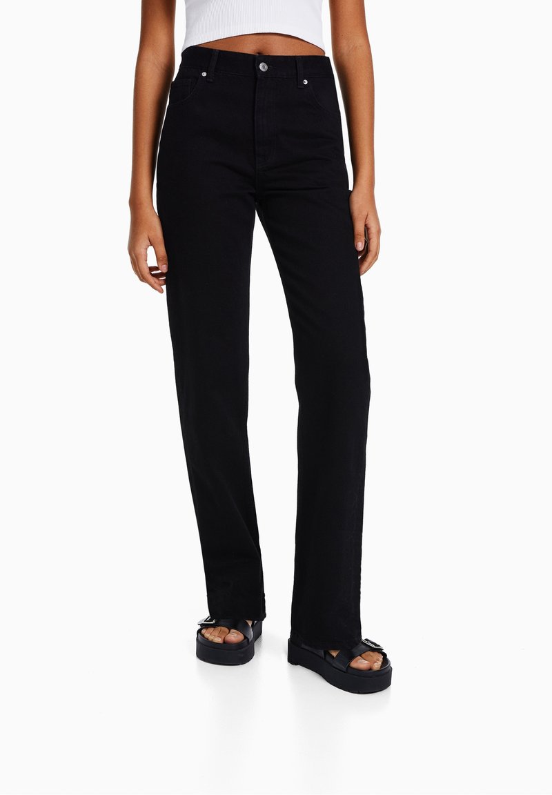 Jeans negros de pierna ancha en tejido resistente, con cierre de botón, diseño de cinco bolsillos y una textura ligeramente descolorida. Combinados con sandalias de plataforma.