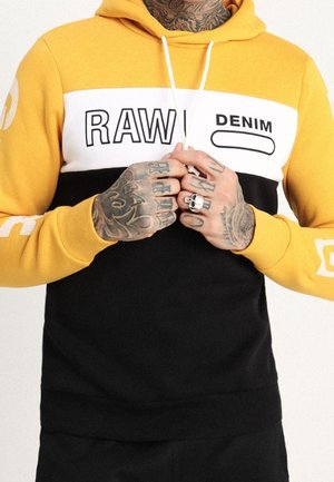 Mężczyzna w żółto-czarnej bluzie z kapturem z napisem "RAW DENIM", pokazujący wytatuowane dłonie i palce z pierścieniami, trzymający sznurki od kaptura.
