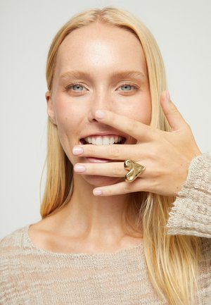 Mujer rubia con ojos azules sonríe, su mano con un gran anillo dorado en forma de corazón cubre su boca, lleva un suéter de punto beige.