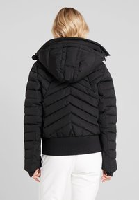 Femme aux cheveux blonds courts portant une veste matelassée à capuche noire et un pantalon blanc, vue de dos sur un fond gris.