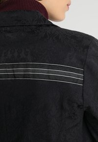 Czarna kurtka z teksturowym kwiatowym wzorem, bordowym ściągaczem przy kołnierzu oraz poziomymi białymi paskami na plecach.