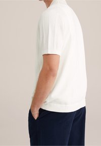 Kortärmad, vit stickad pikétröja med texturerade mönster på axlarna, avslappnad passform, kombinerad med mörkblå byxor.