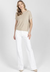 Kobieta ubrana w beżową, krótka dzianinową bluzkę z krótkim rękawem i białe, szerokie spodnie, stojąca na tle jednolitego jasnoszarego tła.