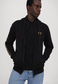 Zwarte hoodie met rits en schaurlkoord, voorzien van een logo en tekst op de mouwen. Draagt over een zwarte polo, gecombineerd met lichtblauwe spijkerbroek.