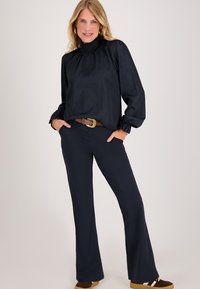 Blonde vrouw staat met haar handen in de zakken, gekleed in een zwarte hoge nek blouse, donkergekleurde broek met een bruine riem en bruine sneakers op een lichte achtergrond.