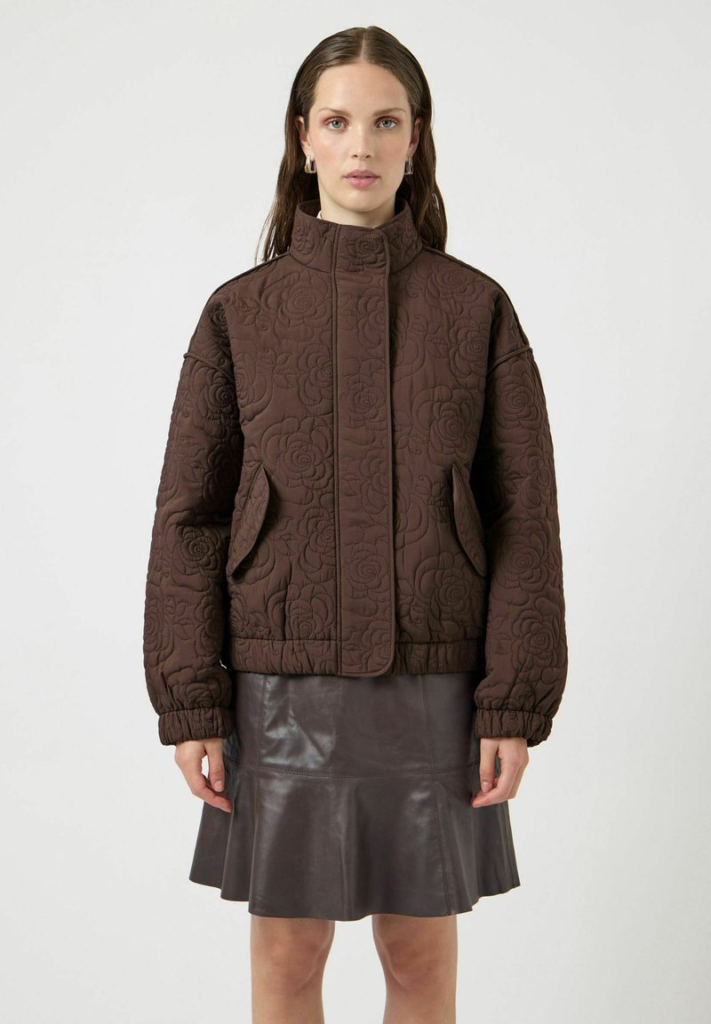 Chaqueta marrón acolchada con textura floral, cuello alto y bolsillos laterales. Se lleva sobre una falda marrón de cuero con vuelo, con puños elásticos.