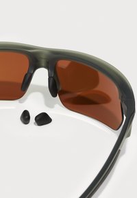 Sonnenbrille mit einem matten olivgrünen Rahmen, braunen Gläsern und Gummi-Ohrpolstern. Enthält zwei zusätzliche schwarze Gummiteile.