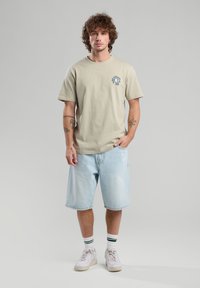 Camiseta de algodón de color beige claro con mangas cortas y un pequeño gráfico en el lado izquierdo del pecho, combinada con pantalones cortos de denim azul y zapatillas blancas.