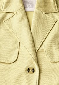 Žlutý blazer s herringbone vzorem a ostrými klopami, který má jednoznačné zapínání na knoflík. Je vyroben z jemně texturované tkaniny a má hladkou podšívku.