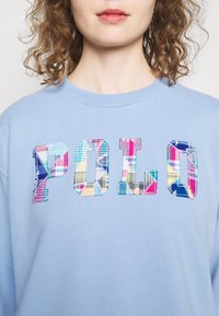 Ljusblå sweatshirt i plyschtyg, med rundad halsringning och flerfärgade, rutigt sydda "POLO"-bokstäver på framsidan.