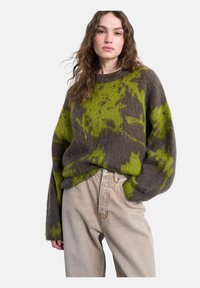Femme aux cheveux ondulés portant un pull ample à motif vert et marron et un pantalon taille haute beige, debout avec une main dans la poche.