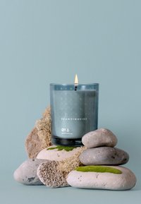 Glass candle with a blue-green wax, flame lit, placed on smooth stones. Candle features embossed patterns and brand name, "SKANDINAVISK."