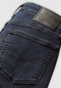 Dark denim jeans featuring a distinctive back pocket shape, brown stitching, and a black leather logo patch. Smooth texture and classic design.