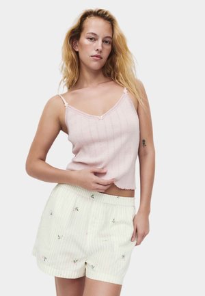 Giovane donna con capelli biondi sciolti che indossa una canottiera rosa chiaro e pantaloncini crema con piccoli motivi floreali su uno sfondo uniforme.