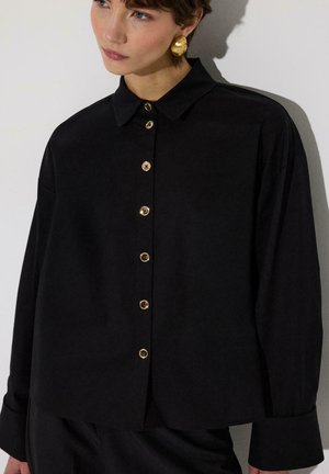 Młoda kobieta w czarnej koszuli zapinanej na guziki z złotymi guzikami i dużymi złotymi kolczykami, patrząca lekko na bok.