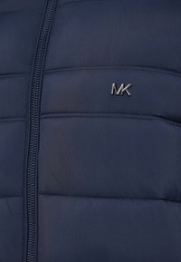Tums navy zils pūkainais jaka ar šuvumiem un sudraba metāla "MK" logotipu kreisajā krūtī. Aprīkots ar priekšējo rāvējslēdzēju un gludu tekstūru.