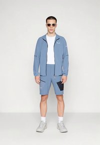 Lys blå atletisk antrekk med en jakke med glidelås og matchende shorts. Shorts har mørkeblå detaljer og lommer. Joggesko fullfører utseendet.