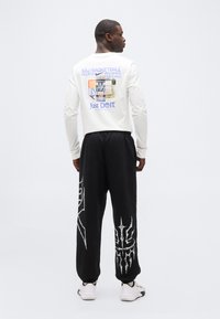 Άνδρας με την πλάτη γυρισμένη, φοράει λευκό μακρυμάνικο μπλουζάκι Nike με εκτύπωση γηπέδου μπάσκετ, μαύρο παντελόνι με γραφικά στα πόδια και λευκά αθλητικά παπούτσια.