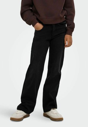 Modèle portant un sweat-shirt marron foncé, un jean noir à jambes larges et des baskets blanches sur un fond uni clair.