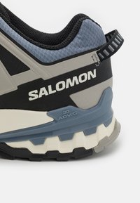 Sive i crne planinarske cipele s teksturiranim gornjim dijelom, plavim naglascima i dubokim potplatom. Imaju povlačnu traku i "SALOMON" logotip.