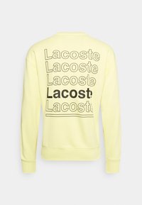 Jasnożółta bluza z powtarzającym się czarnym napisem "Lacoste" na plecach, z jednym pogrubionym "Lacoste" na środku.