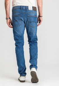 Mężczyzna w obcisłych niebieskich jeansach z metką na pasku, białych tenisówkach i białej koszuli, stojący na jednolitym tle.