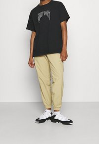 Černé grafické tričko s bílým potiskem, béžové elastické kalhoty a černobílé tenisky se pruhovaným designem a gumovou podrážkou.