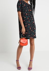 Robe noire à fleurs avec manches courtes, associée à un sac à main orange vif et des sandales à talons violettes, mettant en valeur des couleurs et des motifs vibrants.