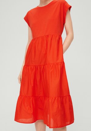 Femme portant une robe midi rouge vif à manches courtes, à volants, avec des coutures froncées et une coupe ample, sur un fond clair uni.