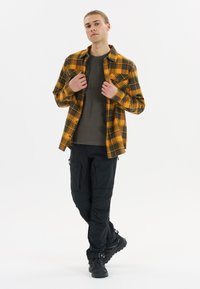 Flanellen overhemd in een geel en zwart geruit patroon over een grijs T-shirt, gecombineerd met zwarte cargobroek en zwarte veterschoenen.