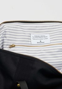 Černá textilní taška se zlatým zipem, uvnitř s pruhovaným šedobílým vzorem a štítkem, který uvádí, že je ručně vyrobená v Německu.