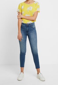 Top corto de manga corta con estampado floral amarillo, combinado con unos jeans slim fit de color azul desvanecido y zapatillas blancas. El conjunto presenta materiales casuales y ligeros.