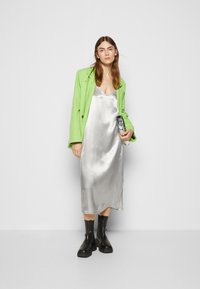 Vestido de seda prateada ajustado com um decote em V profundo, combinado com um blusão oversized verde brilhante. Botas de tornozelo pretas completam o look.